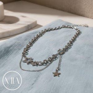 Pode incluir: Um colar prateado com um design em camadas. O colar apresenta uma corrente com pequenas elos redondos e uma seção com três pingentes em forma de estrela. Um único pingente de estrela pende de uma corrente delicada. O colar repousa sobre um tecido azul claro.