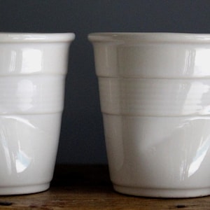 May include: Three white ceramic cups with a glossy finish. Each cup has a cylindrical shape with horizontal banded detailing. The cups are arranged in a row, showcasing their clean, minimalist design. The background is a muted, dark color.