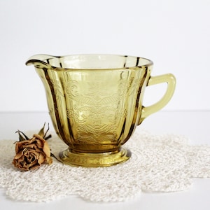 May include: A vintage amber glass creamer with a delicate etched floral design. The creamer has a rounded body and a small handle.