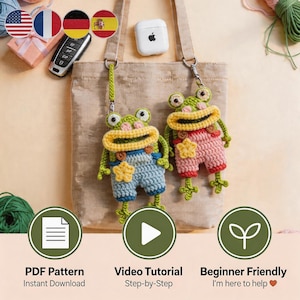 May include: Two handmade crochet frog keychains. One frog wears blue overalls with a yellow star, the other wears pink overalls. The frogs have green bodies, yellow mouths, and large, round eyes. The image also includes text that says "PDF Pattern Instant Download", "Video Tutorial Step-by-Step", and "Beginner Friendly I'm here to help".