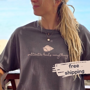 Può includere: T-shirt grigio scuro con una grafica a conchiglia e la frase "saltwater heals everything" in corsivo. La maglietta ha una vestibilità ampia e maniche corte.