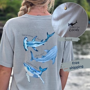 Könnte beinhalten: Hellgraues T-Shirt mit Aquarellillustrationen eines Hammerhais, Walhais und Buckelwals. Der Text "Respect the Locals" ist auf der Vorderseite aufgedruckt. Das Shirt hat eine lockere Passform und kurze Ärmel.