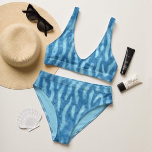 Könnte beinhalten: Blaues Bikini-Set mit einzigartigem Muster, bestehend aus einem Oberteil mit V-Ausschnitt und einem hoch taillierten Unterteil. Zubehör sind ein Sonnenhut, Sonnenbrille, Sonnencreme und eine Muschel.