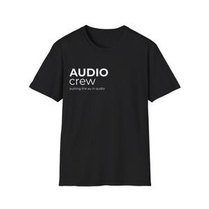 Op de afbeelding: Zwart T-shirt met ronde hals en de witte woorden "AUDIO crew". Daaronder staat de zin "putting the au in audio" in een kleiner lettertype. Het shirt is gemaakt van een zacht materiaal.