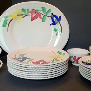 May include: A ceramic dinnerware set featuring a white base with hand-painted floral motifs in red, yellow, and blue with green leaves. The collection includes a large serving platter, several plates, saucers, and teacups, all with a beaded edge.