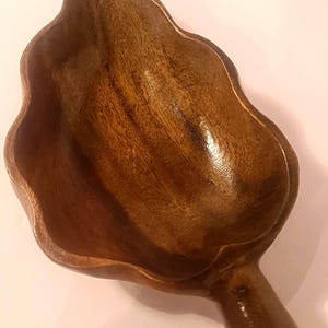 May include: A dark brown wooden bowl carved into the shape of a leaf with a smooth, polished finish. The bowl features a deep, concave center and a short, cylindrical stem at the base, showcasing the natural wood grain patterns throughout the surface.