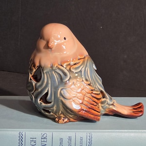 May include: A ceramic bird figurine featuring a glossy finish with blended peach, blue, and brown tones. The body has intricate cut-out patterns and sculpted feather details. It is positioned on a light blue book titled High Towers.