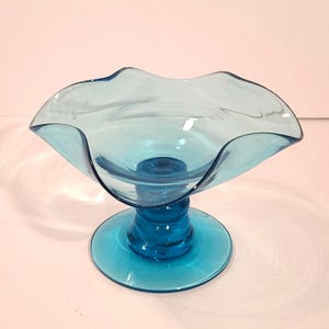 May include: A translucent turquoise glass compote dish featuring a wide, ruffled, scalloped rim and a smooth, circular pedestal base. The bowl transitions from a lighter, pale blue at the top edge to a deeper, saturated blue at the base.