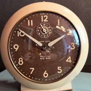 Pode incluir: Um despertador redondo Westclox Big Ben com uma caixa de metal na cor creme e um mostrador castanho escuro. O relógio apresenta algarismos arábicos brancos, ponteiros brancos e um mostrador secundário circular para o alarme no centro.