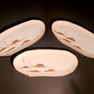 Puede incluir: Tres platos ovalados de cerámica con una base de color crema claro y una sutil textura circular concéntrica. Cada plato presenta un diseño botánico pintado en marrón de juncos y hojas en un lado, con un borde marrón a juego.