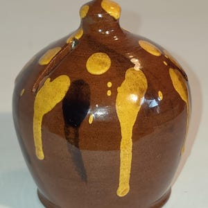 May include: A small, rounded ceramic jug with a glossy dark brown glaze. The surface features decorative yellow drips and spots that run vertically from the narrow neck down the sides of the body, creating a textured, layered appearance.