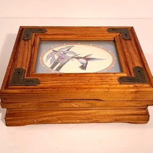 May include: A square wooden box with a natural grain finish, featuring dark metal corner accents. The lid has a glass-covered inset displaying a circular illustration of a hummingbird hovering near a purple flower against a light blue background.
