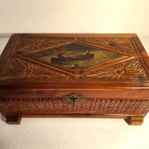 May include: A rectangular wooden box with a hinged lid featuring a central diamond-shaped panel depicting a painted scene of a person in a canoe on water. The lid and sides are decorated with carved floral and leaf patterns and a metal latch.