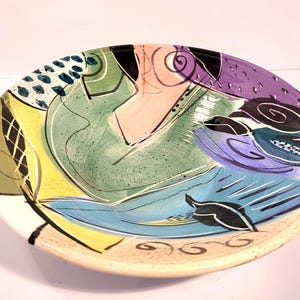 May include: A round ceramic bowl featuring a hand-painted abstract design with vibrant splashes of green, yellow, purple, and blue. The surface displays black brushstrokes, swirling lines, and dotted patterns over a speckled cream-colored base.