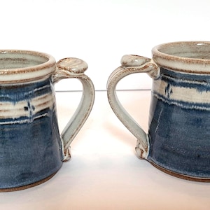 May include: Two ceramic mugs featuring a blue and cream glaze with a horizontal band pattern. Each mug has a sturdy handle with a thumb rest at the top and a visible brown clay base. The exterior surface displays a mottled blue finish.