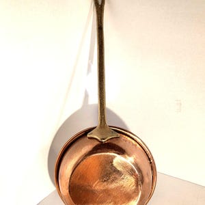 Op de afbeelding: Een ronde koperen pan met een gepolijste metalen afwerking en een lange, getextureerde messing handgreep bevestigd aan een driehoekige basis. De pan heeft een glad, reflecterend oppervlak en een stevige, langwerpige handgreep met een gat.