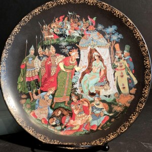 Op de afbeelding: Een ronde zwarte decoratieve schaal met een gedetailleerde, kleurrijke illustratie van een koninklijk hofscène met figuren in historische kleding, waaronder soldaten, een koning en een zittend persoon onder een baldakijn, met gouden rand.