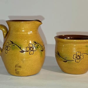 May include: A yellow ceramic pitcher and a matching cup, both featuring a dark brown interior and a hand-painted vine pattern with small flowers in dark brown and green accents. The pitcher has a curved handle and a pouring spout.