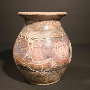 May include: A round ceramic vase with a mottled brown and purple finish. The surface features etched white line drawings of a flute-playing figure, a sun, and a spiral design. The vase has a wide, flared rim and a smooth, rounded body shape.