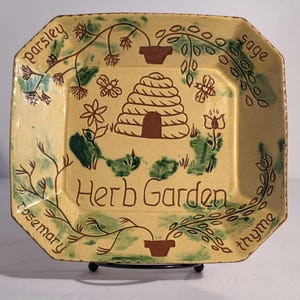 May include: A square ceramic plate with a cream-colored glaze and brown hand-painted details. The center features a beehive, bees, and flowers. The text "Herb Garden" is written in the center, surrounded by the words "parsley," "sage," "rosemary," and "thyme."