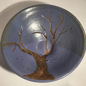 May include: A round ceramic bowl with a smooth, glossy periwinkle blue glaze. The interior features a detailed, textured brown tree design with bare branches extending upwards. The surface has subtle dark speckles throughout the blue finish.