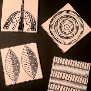 May include: Four square ceramic coasters with white surfaces and black ink illustrations. Designs include a central arch, a diamond-shaped radial pattern, two leaf-like shapes with cross-hatching, and a grid of vertical and horizontal lines.