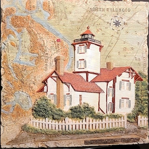 May include: A three-dimensional wall plaque featuring a relief of the North Wildwood lighthouse. The scene includes a nautical map background, a white house with a red roof, a brick chimney, and a white picket fence, signed Donna Elias.