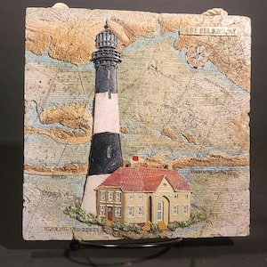 Könnte beinhalten: Eine quadratische Wandplatte mit einer Reliefkarte von Fire Island, NY, einem markanten schwarz-weißen Leuchtturm und einem beigen Haus mit rotem Dach. Die Karte enthält nautische Gitterlinien, eine Windrose und den Text Fire Island, NY.