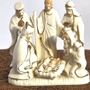May include: A porcelain nativity figurine set featuring a central standing figure, a kneeling figure, a standing figure with a staff, and a figure holding a gift, all surrounding a small infant lying in a manger, finished in cream with gold trim.