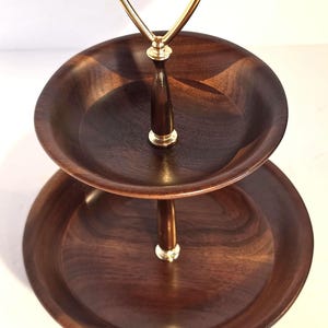 May include: A two-tiered serving stand featuring two circular dark brown wooden trays connected by a central metal post. The stand is topped with a gold-colored metal handle, and the wood grain is visible across the surface of both trays.