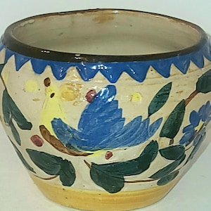 Peut inclure: Un pot de fleurs en céramique ronde avec une base couleur crème, orné d'un motif peint à la main représentant un oiseau jaune et bleu perché sur une branche. Le bord présente un motif en zigzag bleu et la base est soulignée d'une bande jaune.
