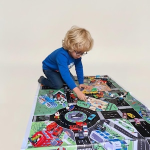Puede incluir: Un niño juega con coches de juguete sobre una colorida alfombra de juego. La alfombra presenta un diseño de mapa de carreteras con calles, edificios y una estación de bomberos. Varios coches de juguete rojos, azules, amarillos y verdes están esparcidos.