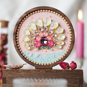 Könnte beinhalten: Ein runder Holzrahmen mit einem dekorativen Muschelmotiv aus irisierenden, rosa und violetten facettierten Kristallen auf hellrosa Stoffhintergrund, ergänzt durch hellblaue, bogenförmige Akzente und einen Rand aus kleinen weißen Perlen.