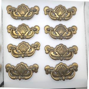 Puede incluir: Un juego de ocho tiradores de cajón de latón a juego dispuestos en dos columnas. Cada tirador cuenta con una placa decorativa con un motivo central de concha y un asa curva y oscilante unida a dos puntos de montaje laterales.