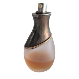 May include: A perfume bottle with a frosted glass base containing a peach-colored liquid. The upper portion is a metallic silver-toned cap with a diagonal cut, separated from the glass by a thin gold-toned ring. A silver spray nozzle is visible.