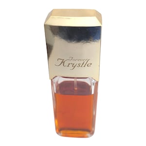 May include: A rectangular glass perfume bottle filled with amber-colored liquid, featuring a tall, reflective gold-toned cap. The front of the gold cap is embossed with the text Forever Krystle in a script font, and a clear spray nozzle is visible.