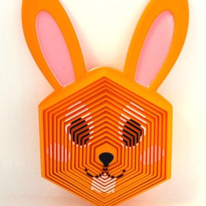 May include: A layered, orange geometric bunny head with pink inner ears and black eyes. The bunny's face is a series of hexagonal shapes, creating a 3D effect. The design includes a small black nose and a white and black pattern for the mouth.