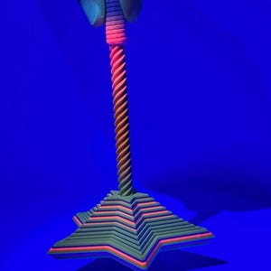 Könnte beinhalten: Ein neonfarbenes, sternförmiges Objekt mit einem gedrehten Stiel, das von einer Hand gehalten wird. Der Stern hat geschichtete Kanten in Grün, Orange und Rosa. Der Stiel ist orange und gelb. Der Hintergrund ist einfarbig blau.