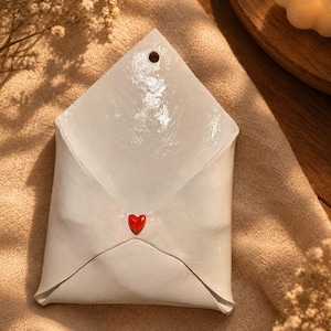 May include: A white ceramic envelope-shaped wall hanging with a glossy finish. The envelope features a small, bright red heart detail at the centre of the flap and a single circular hole at the top point for hanging purposes on a wall or surface.