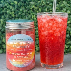 Op de afbeelding: Een glas rode drank met ijs en een rietje staat naast een pot Beachfront Property energy tea mix. Het etiket op de pot luidt "Beachfront Property" en geeft smaken aan. De drank is kersen-, ananas- en pina colada-smaak.