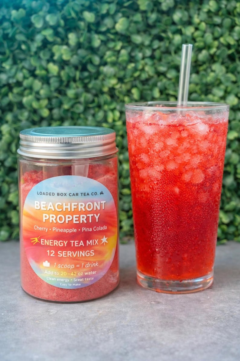 Op de afbeelding: Een glas rode drank met ijs en een rietje staat naast een pot Beachfront Property energy tea mix. Het etiket op de pot luidt "Beachfront Property" en geeft smaken aan. De drank is kersen-, ananas- en pina colada-smaak.