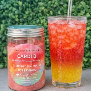 Op de afbeelding: Een potje "Cardi B" energy theemix naast een glas van de gemengde drank. De pot heeft een zilveren deksel en een label met de naam van de thee en de smaken: watermeloen, ananas en cranberry. De drank is gelaagd met rode, oranje en gele tinten.