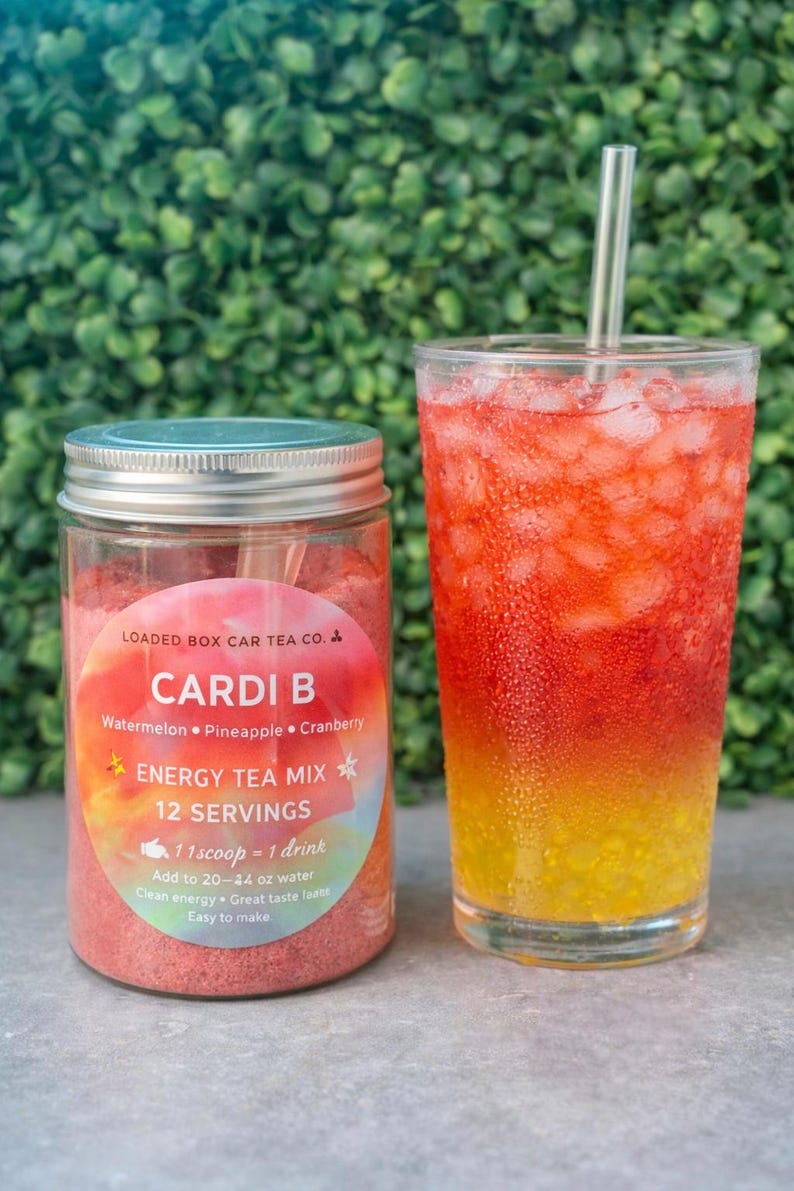 Op de afbeelding: Een potje "Cardi B" energy theemix naast een glas van de gemengde drank. De pot heeft een zilveren deksel en een label met de naam van de thee en de smaken: watermeloen, ananas en cranberry. De drank is gelaagd met rode, oranje en gele tinten.