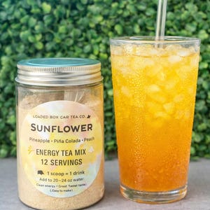 Op de afbeelding: Een glazen pot met Sunflower Energy Tea Mix naast een glas ijsthee. De pot heeft een etiket met de tekst "Sunflower Energy Tea Mix" en smaakdetails. De ijsthee is helder oranje van kleur met ijs en een rietje.