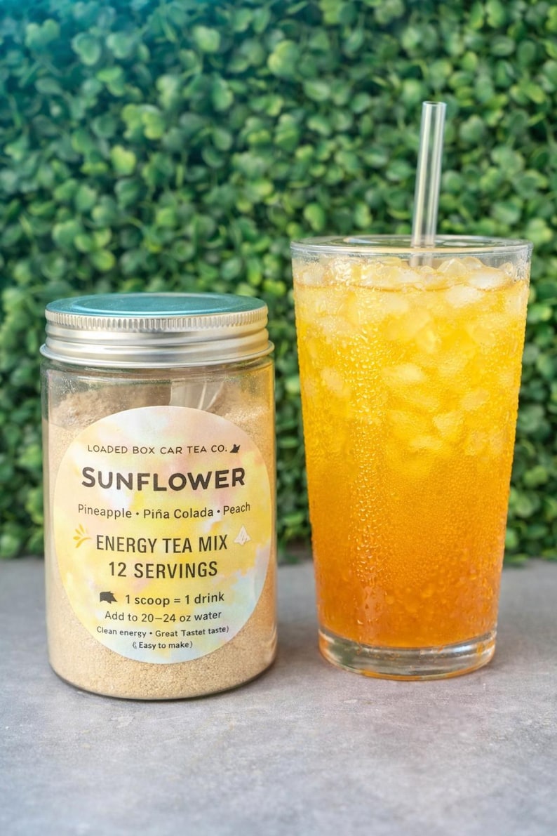 Op de afbeelding: Een glazen pot met Sunflower Energy Tea Mix naast een glas ijsthee. De pot heeft een etiket met de tekst "Sunflower Energy Tea Mix" en smaakdetails. De ijsthee is helder oranje van kleur met ijs en een rietje.