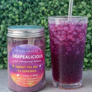 Op de afbeelding: Een pot "Grapealicious" energy tea mix naast een glas paarse drank met ijs en een rietje. Het etiket op de pot vermeldt de smaken druif, granaatappel en framboos. De tekst op de pot zegt "12 porties" en "1 schep = 1 drankje".