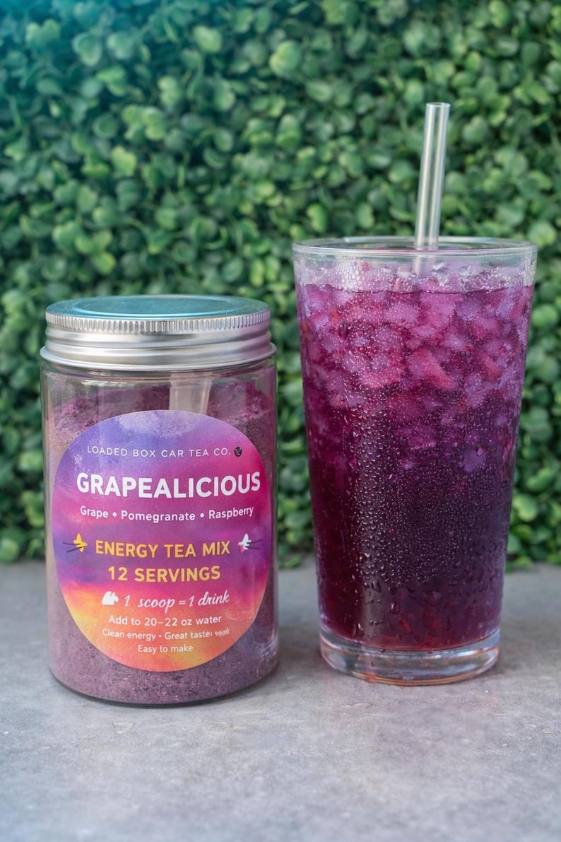 Op de afbeelding: Een pot "Grapealicious" energy tea mix naast een glas paarse drank met ijs en een rietje. Het etiket op de pot vermeldt de smaken druif, granaatappel en framboos. De tekst op de pot zegt "12 porties" en "1 schep = 1 drankje".