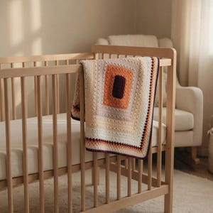 May include: A rectangular crochet blanket featuring a concentric square pattern in shades of cream, beige, light orange, and deep burgundy. The blanket is draped over the wooden railing of a crib, showcasing a textured stitch design throughout.