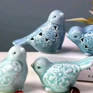 May include: Four ceramic bird figurines in shades of blue and teal. The birds have detailed wing and body patterns, with some featuring cut-out designs. The figurines are arranged on a white surface, creating a decorative display.