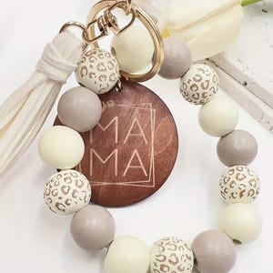 May include: A circular keychain featuring a gold-toned metal ring, a white faux suede tassel, and a series of wooden beads in cream, grey, and leopard print. A central round wooden disc is engraved with the word MAMA inside a square frame.